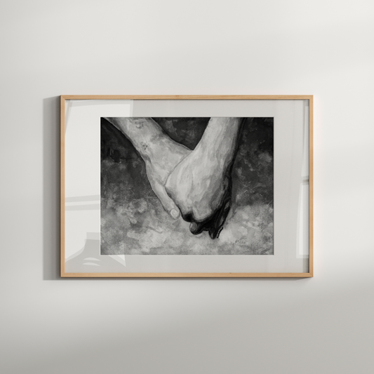 Framed black and white photograph of two hands holding each other on a light gray wall.