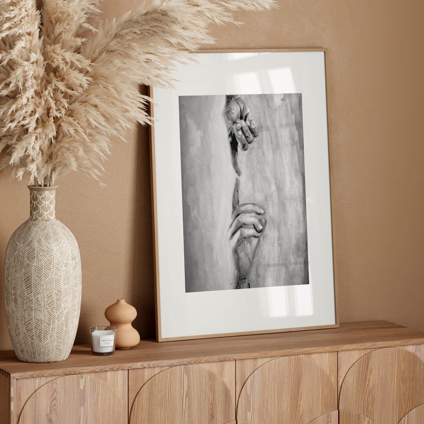 Decorative setup with a vase, candle, and framed artwork on a wooden console against a beige wall.