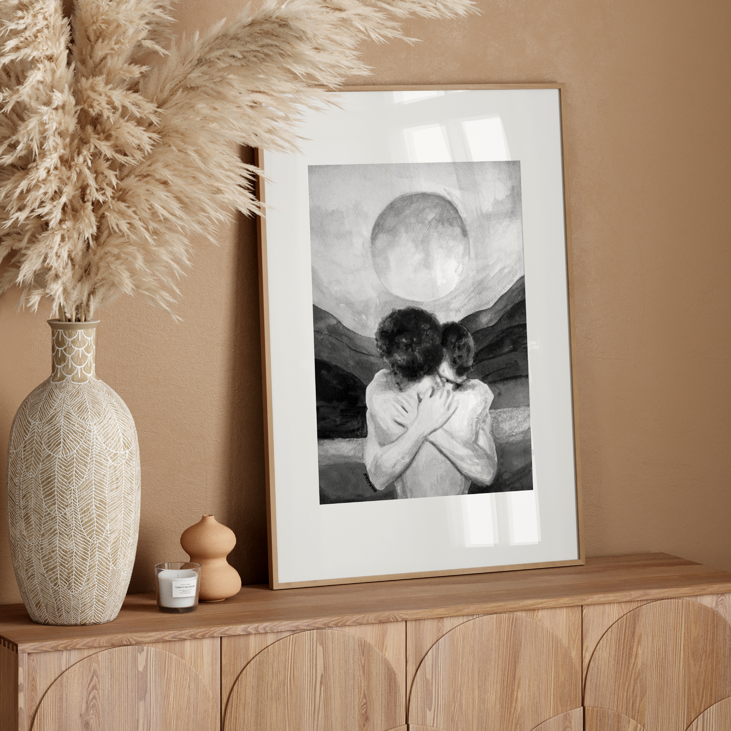 Framed artwork on a wooden console table with decorative vase and pampas grass against a beige wall.