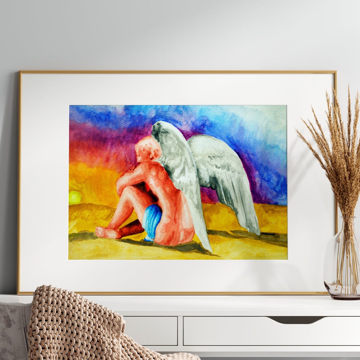 A watercolor painting of an angel sitting next to a sunset, framed in a light wood frame.