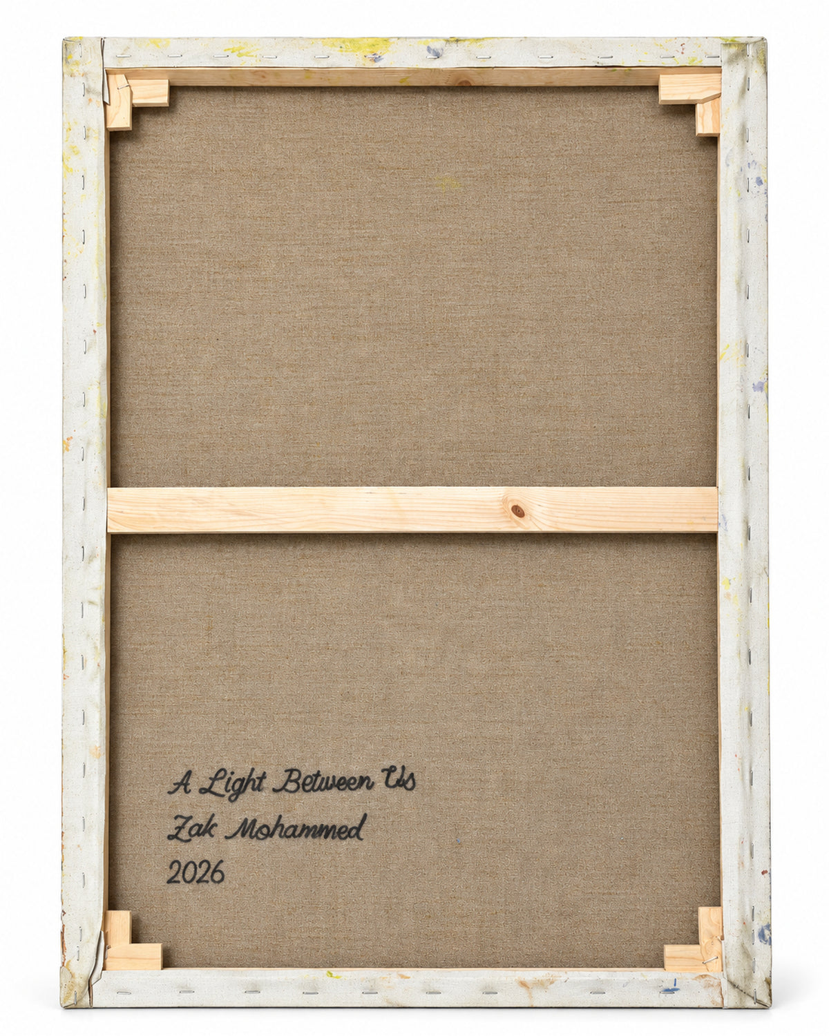 The back of a framed canvas print showing the wooden stretcher bars and corner supports, with the text 'A Light Between Us, Zak Mohammed 2026' printed on the brown linen backing.