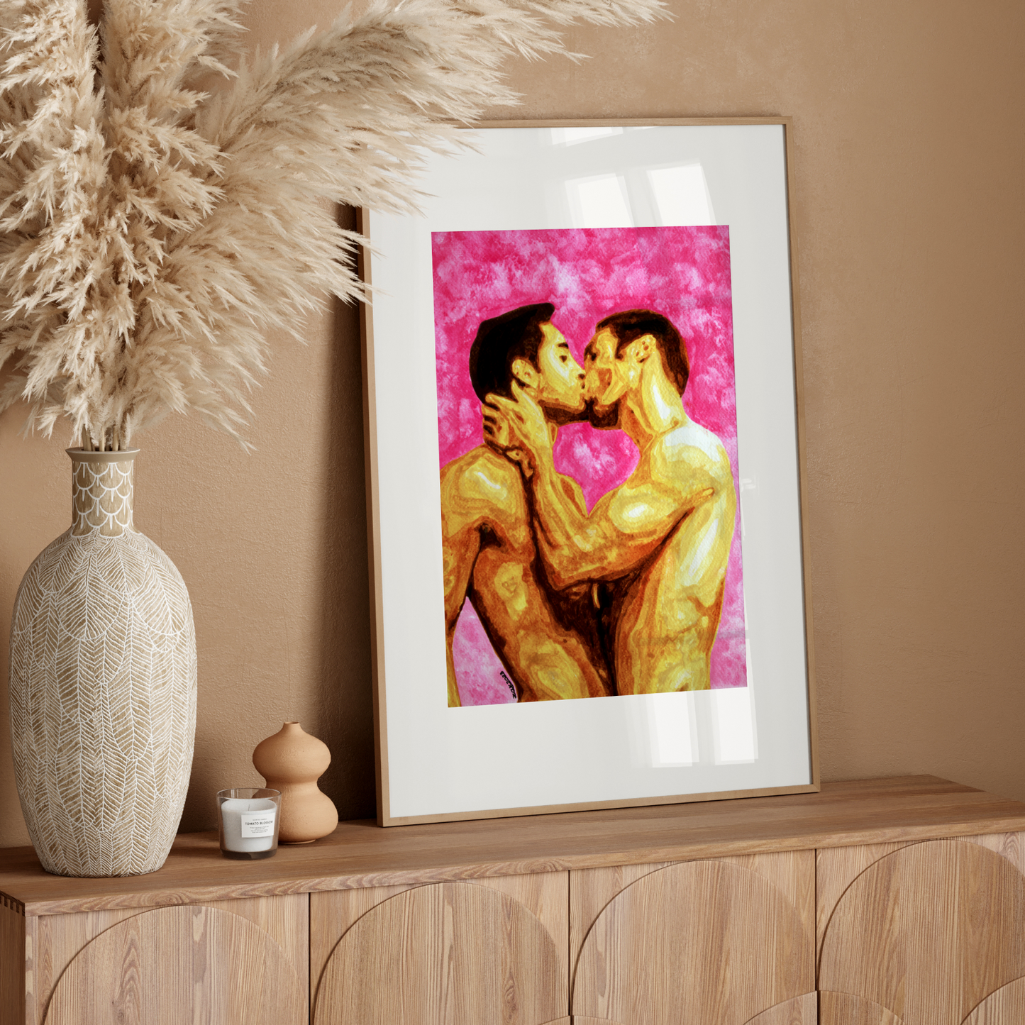 Framed artwork of two people kissing on a pink background, placed on a wooden surface with decorative items.