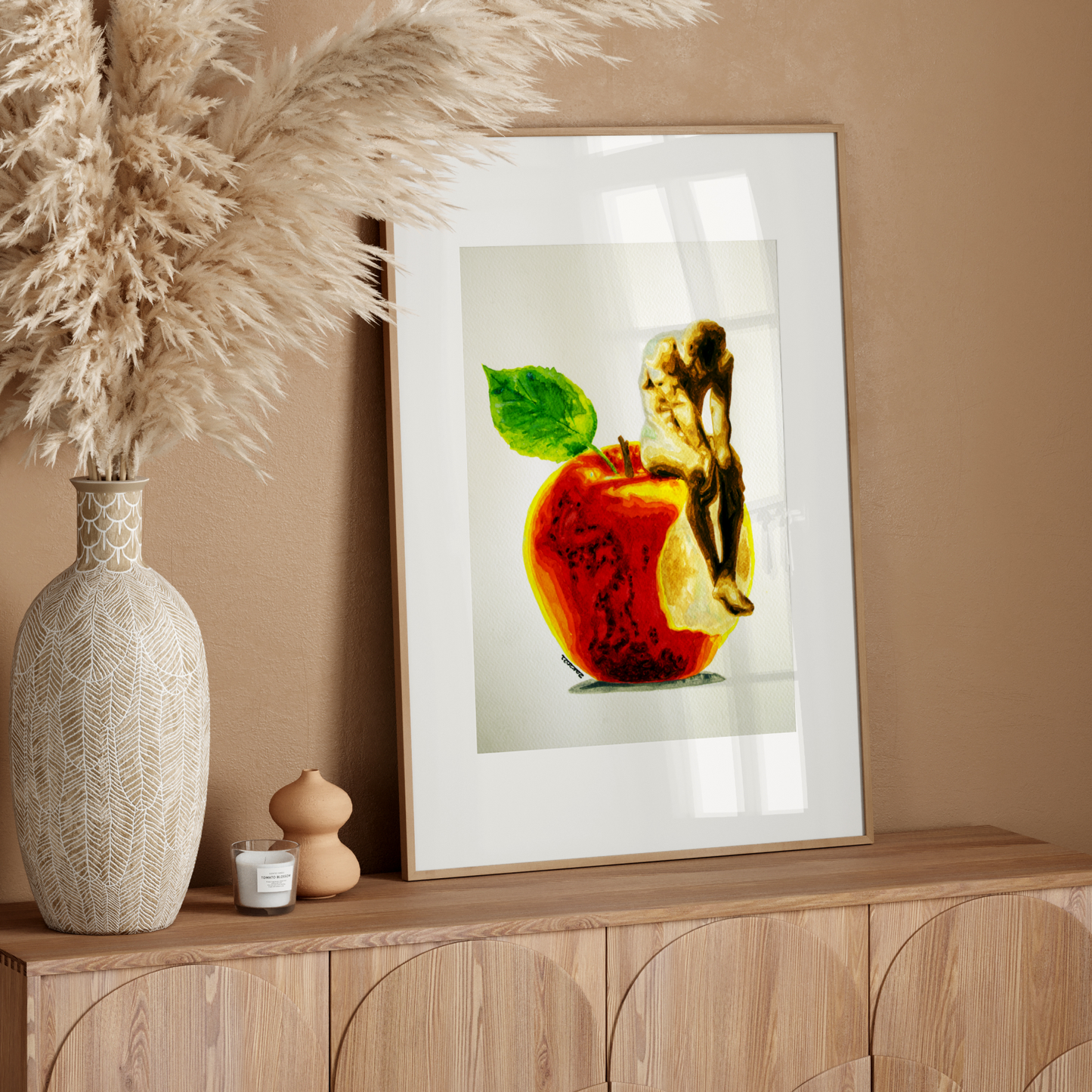 Framed artwork of an apple with a leaf on a wooden sideboard against a beige wall.