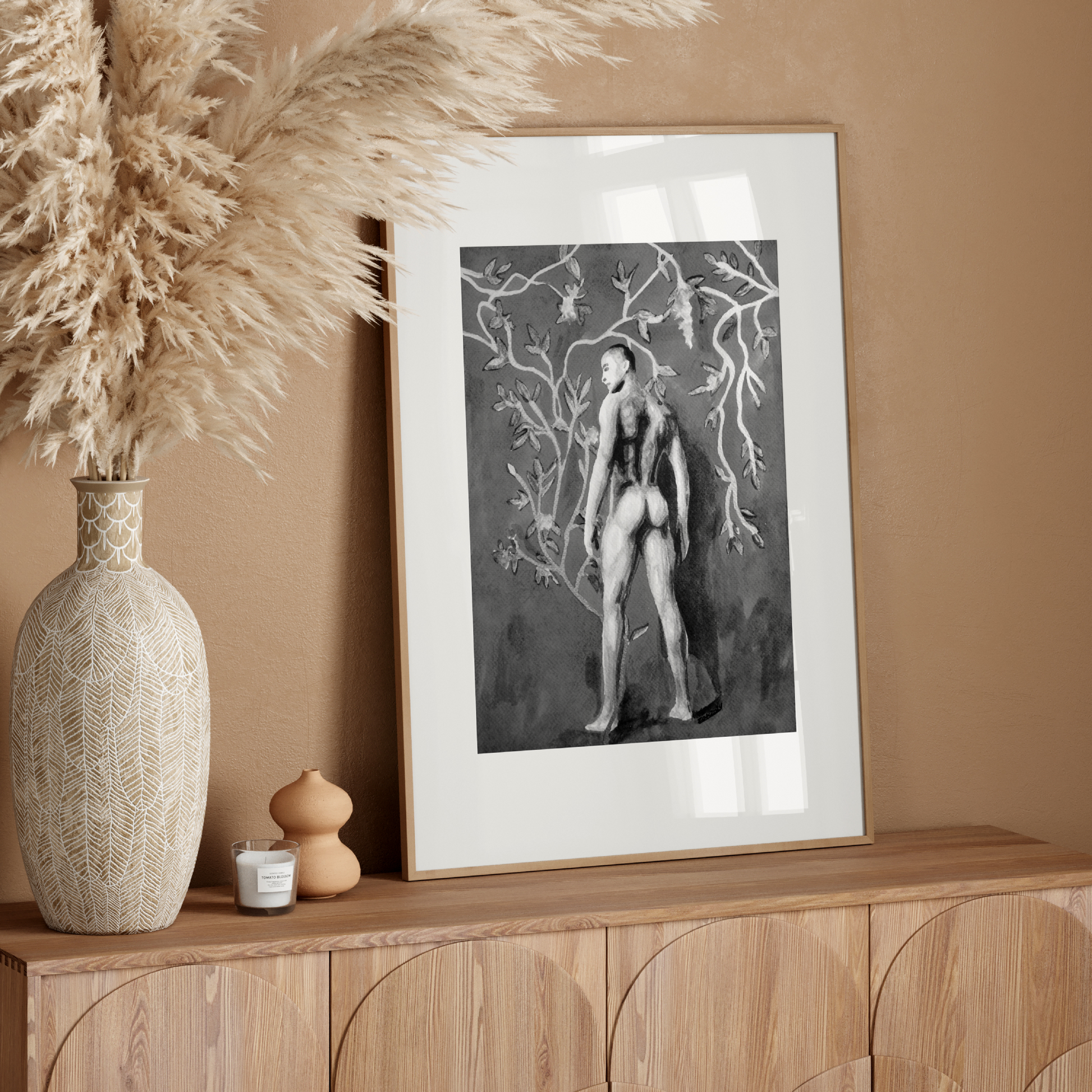 Framed artwork on a wooden console table with decorative vase and pampas grass against a beige wall.