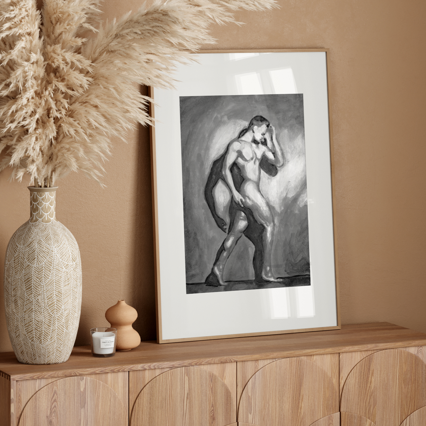 Framed artwork on a wooden console table with decorative vase and pampas grass against a beige wall.