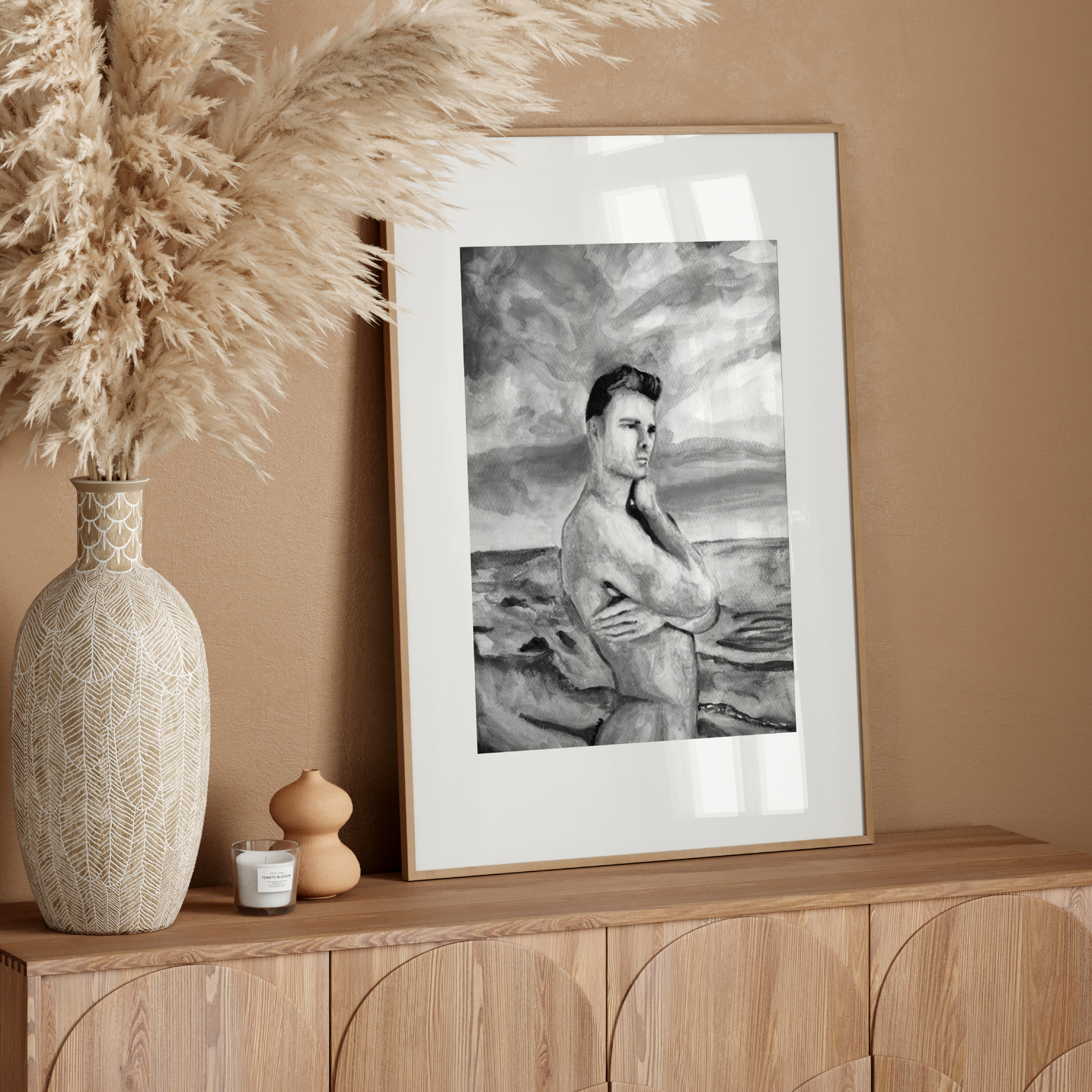 Framed artwork on a wooden console table with decorative vase and pampas grass against a beige wall.