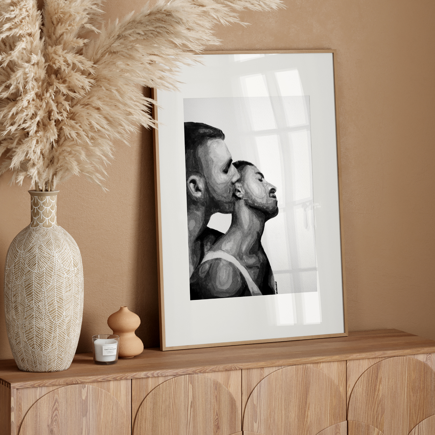 Framed black and white artwork of two men in a close embrace on a white wall.
