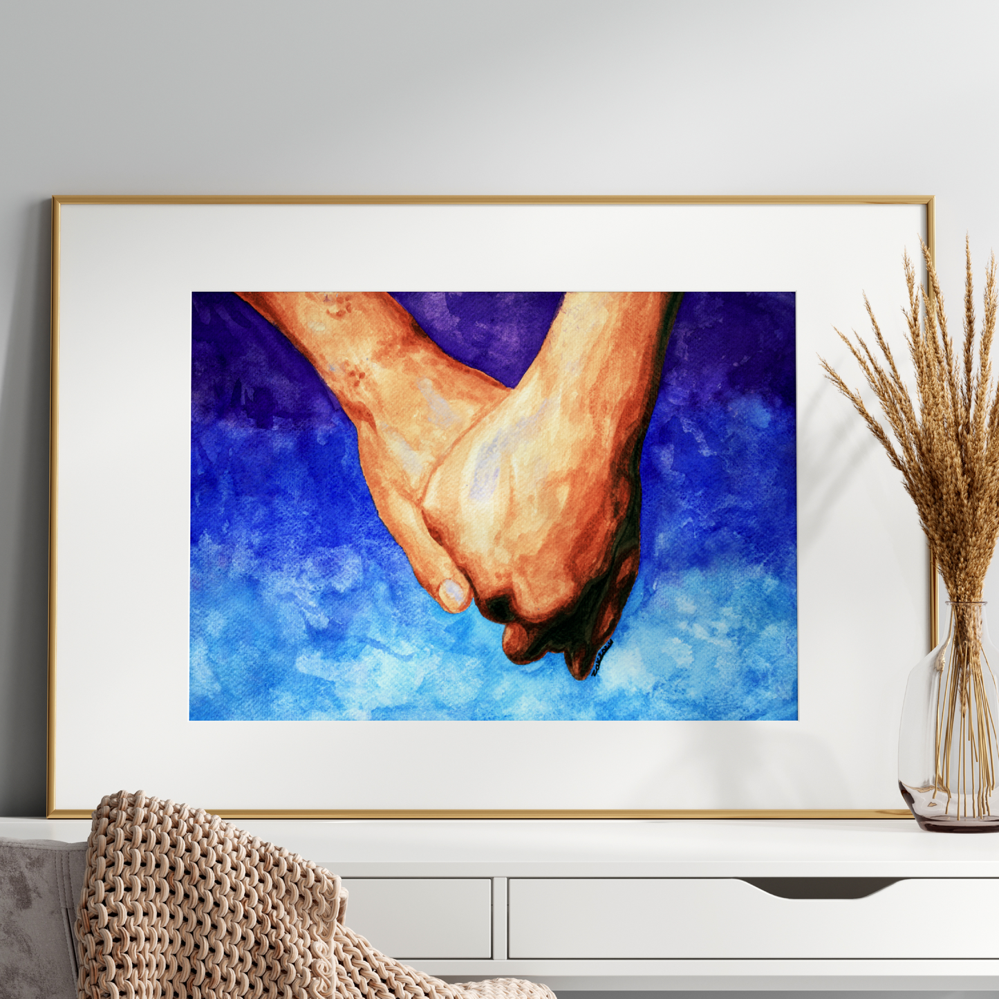 Framed artwork of two hands holding each other on a blue background, displayed on a white wall above a white cabinet.