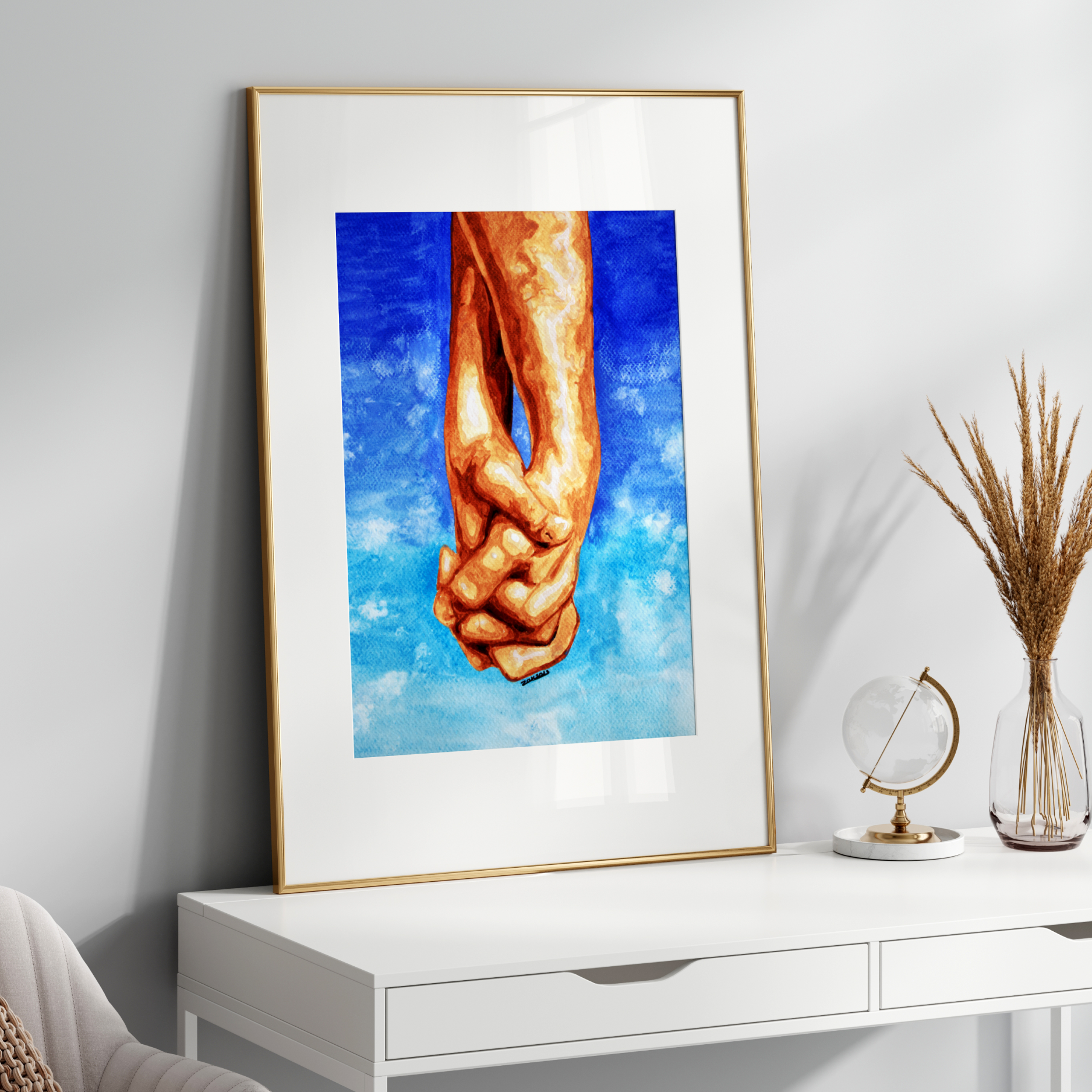 Framed artwork of two hands holding each other against a blue sky with clouds on a white wall.