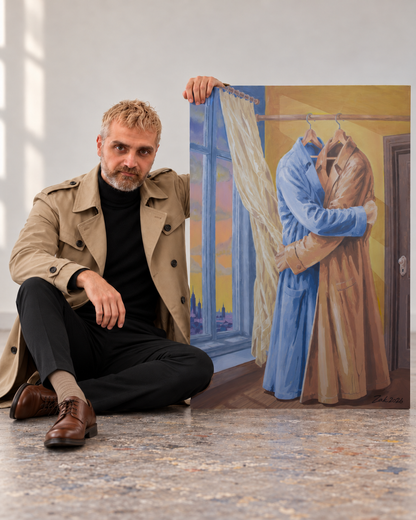 A man in a beige trench coat and black outfit sits on a textured floor next to a large canvas painting depicting two people hugging under a window, with one figure in a blue robe and the other in a tan robe.