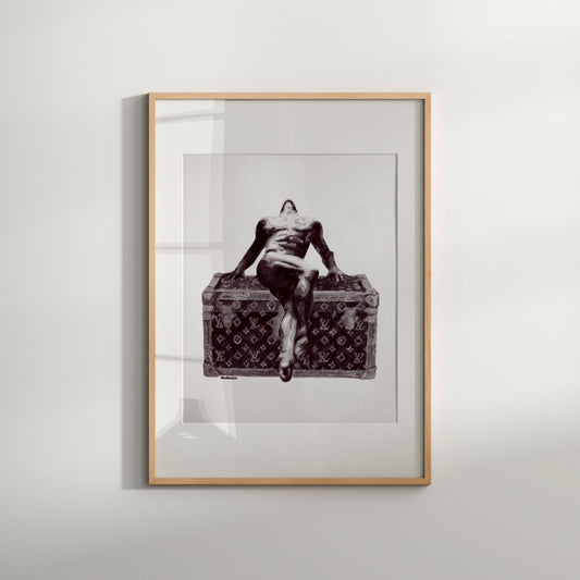 The image depicts a framed black and white print of a nude male figure sitting on a surface, with the figure's hands resting on the object. The artwork is presented in a simple, elegant frame with a white matte border, enhancing its modern aesthetic.
